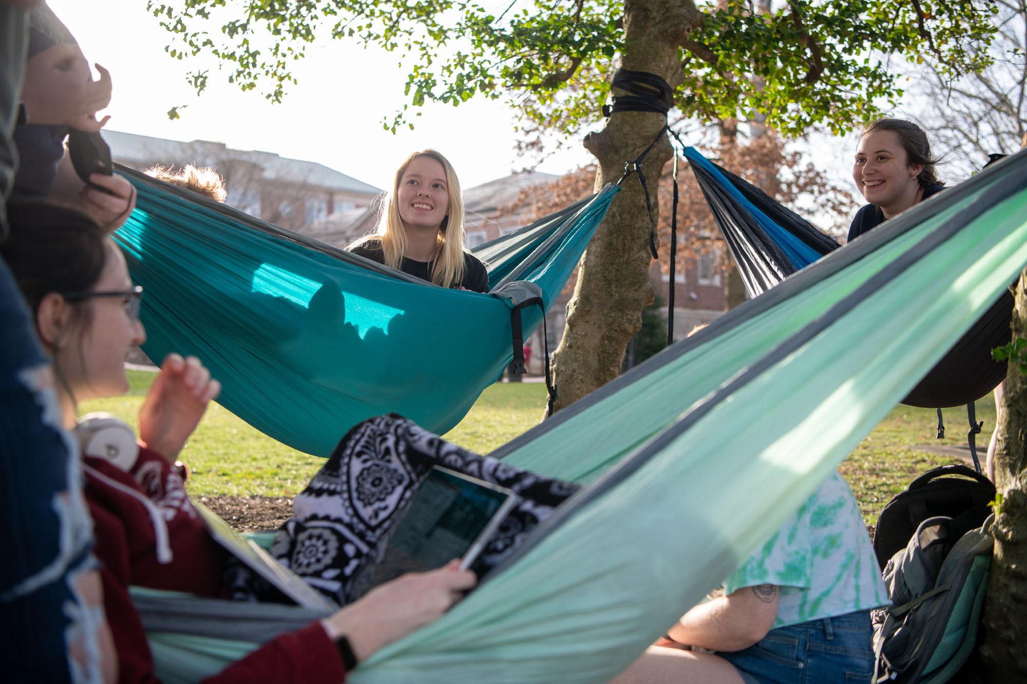 College students smile at each other while they sit in hammocks outdoors